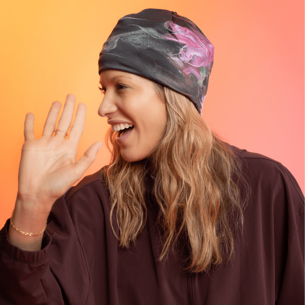 A woman wearing a black beanie with a pink and green floral art print smiles and waves.