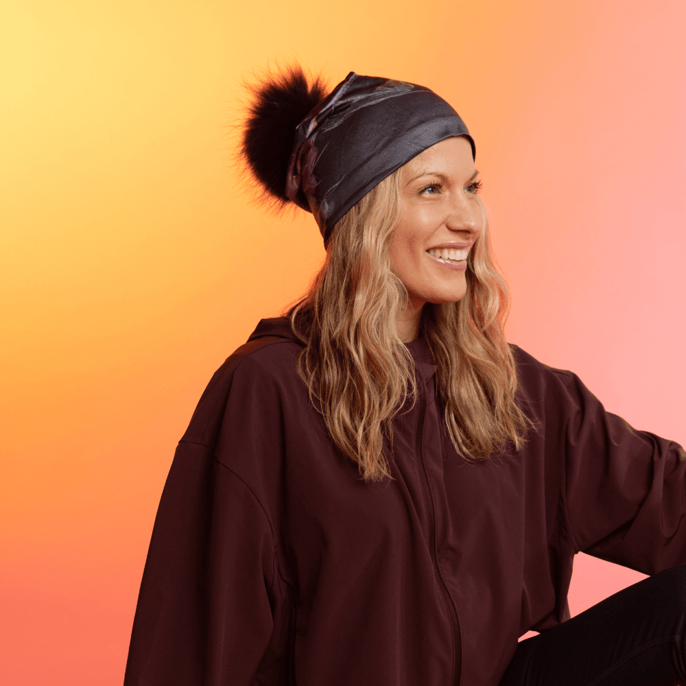 Woman wearing a dark grey beanie with a swirling grey pattern and a black pompom, looking to the side against a gradient background.