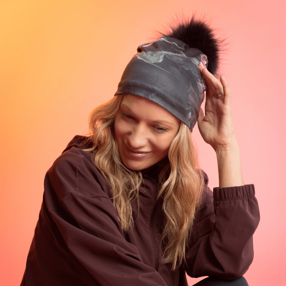 Woman wearing a dark grey beanie with a swirling grey pattern and a black pompom, smiling against a gradient background.