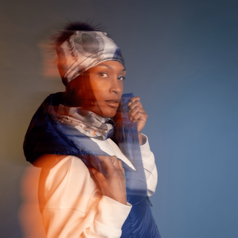 A woman wearing a blue puffer vest over a cream sweatshirt, with a patterned headscarf and neck tube.