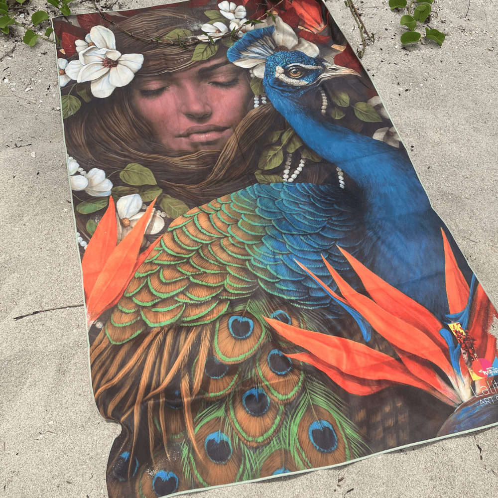 La serviette de plage artistique Chrysalide étendue sur une plage sablonneuse, montrant son art vibrant de paon et de fleurs.