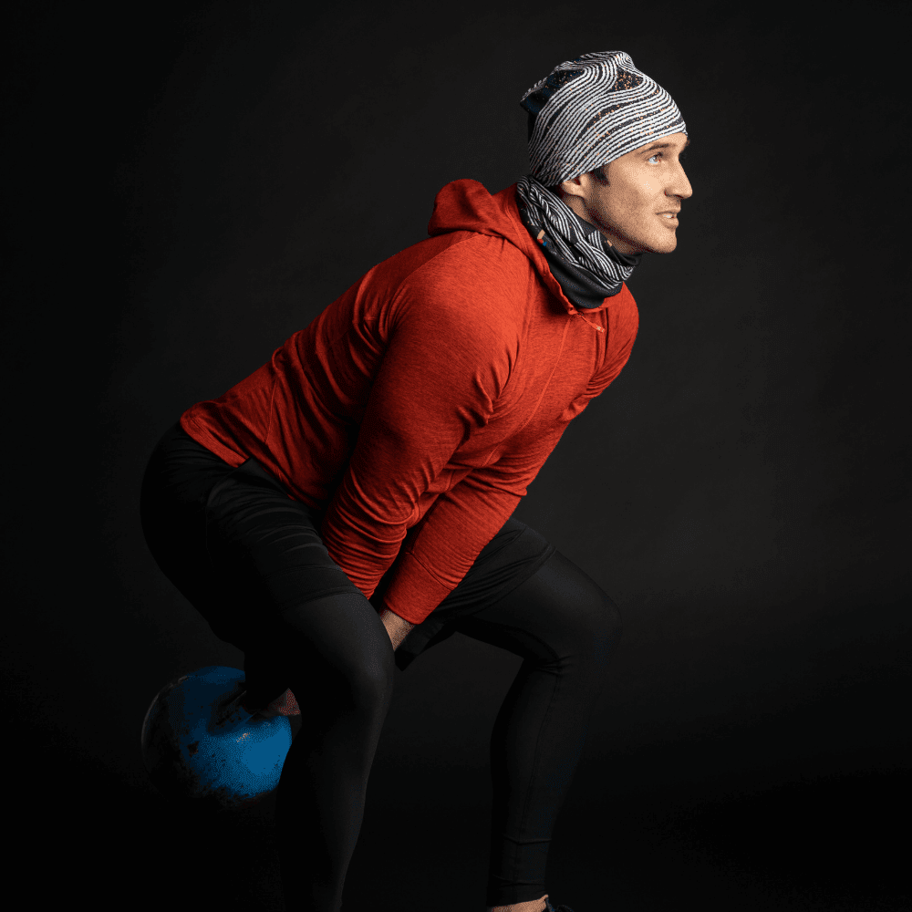 Un homme en sweat à capuche rouge portant une tuque et un cache-cou Dark Steel, effectuant un balancement de kettlebell.