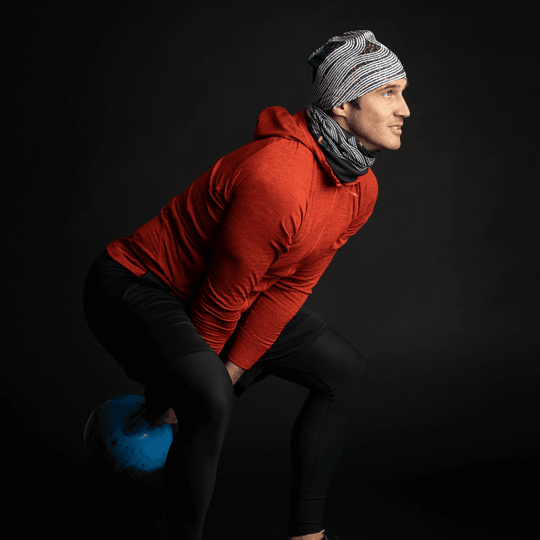 Un homme en sweat à capuche rouge portant une tuque et un cache-cou Dark Steel, effectuant un balancement de kettlebell.
