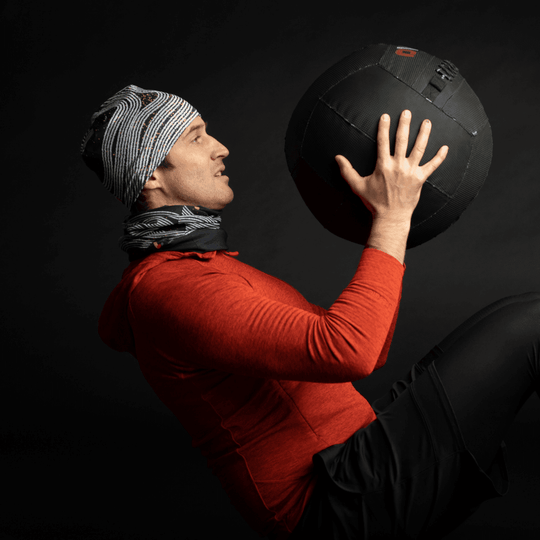 Un homme en sweat à capuche rouge portant une tuque et un cache-cou Dark Steel, tenant un gros ballon médecine.
