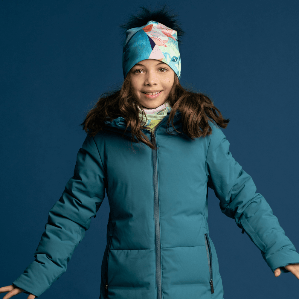 Jeune fille portant une tuque et un cache-cou assortis, habillée d'une veste d'hiver émeraude, souriant sur fond bleu.