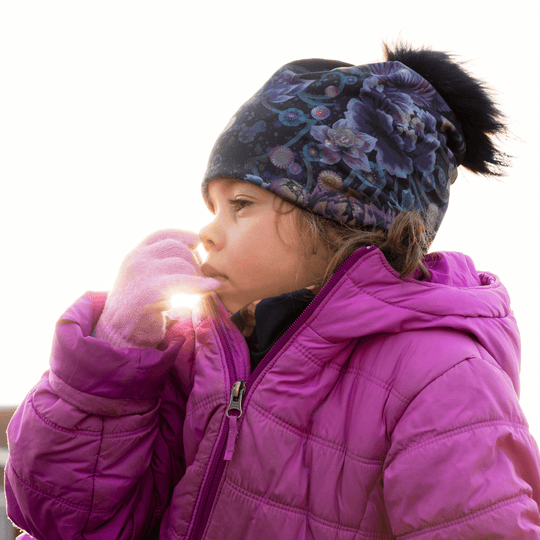 Jeune enfant portant la Tuque Pompon Flora et une veste rose, avec le soleil brillant derrière.