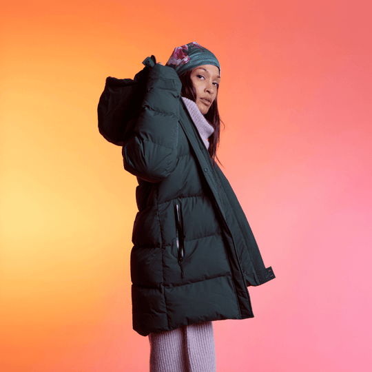 A woman in a winter coat with a patterned art beanie pulled up, looking over her shoulder against a gradient background.