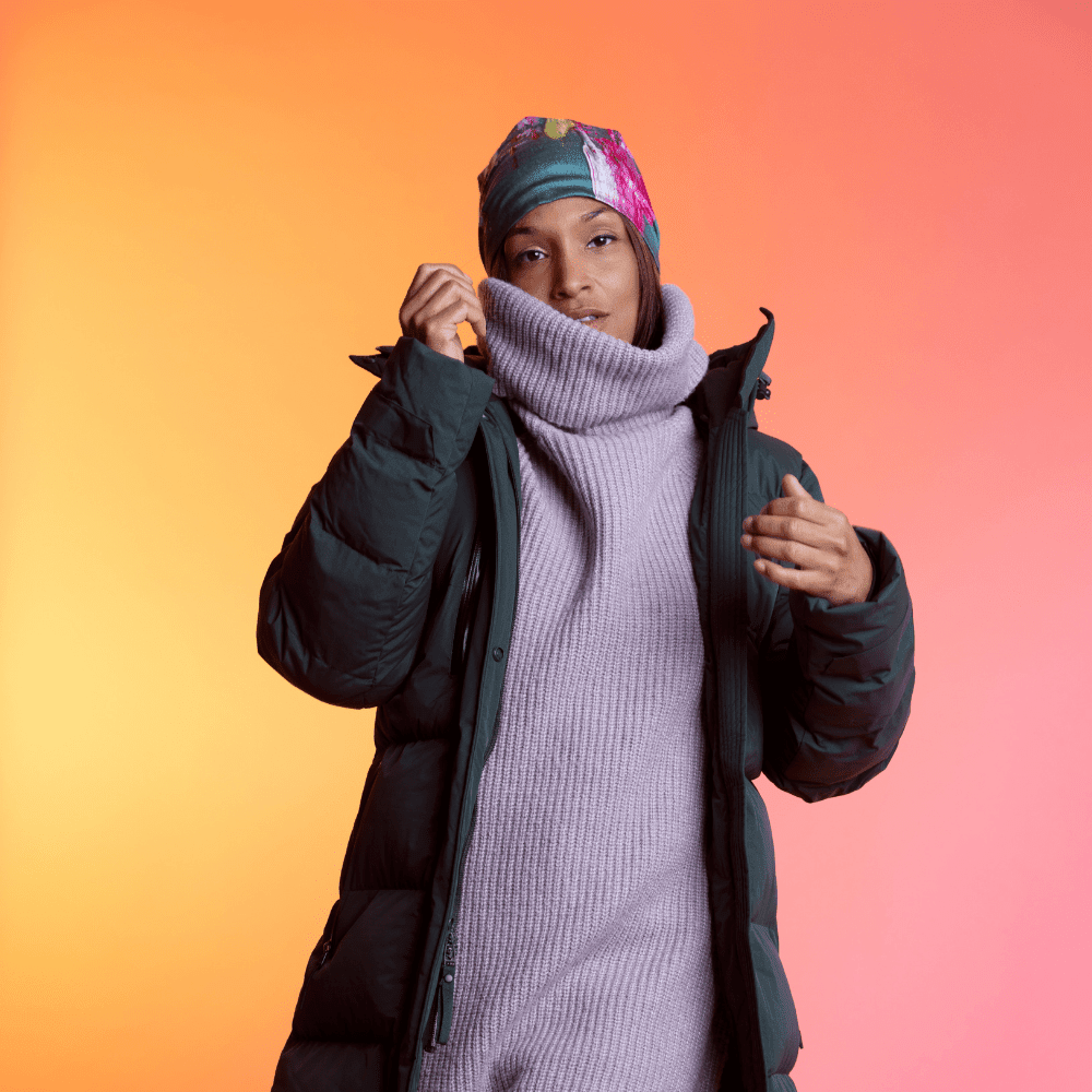 A woman wearing a dark green puffer coat and a light grey turtleneck, adjusting a colourful art print beanie.