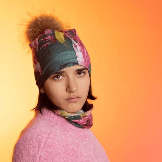 A young person wearing the Flower Glitch Beanie With Pompom and a matching neck gaiter, looking forward against a warm gradient background.