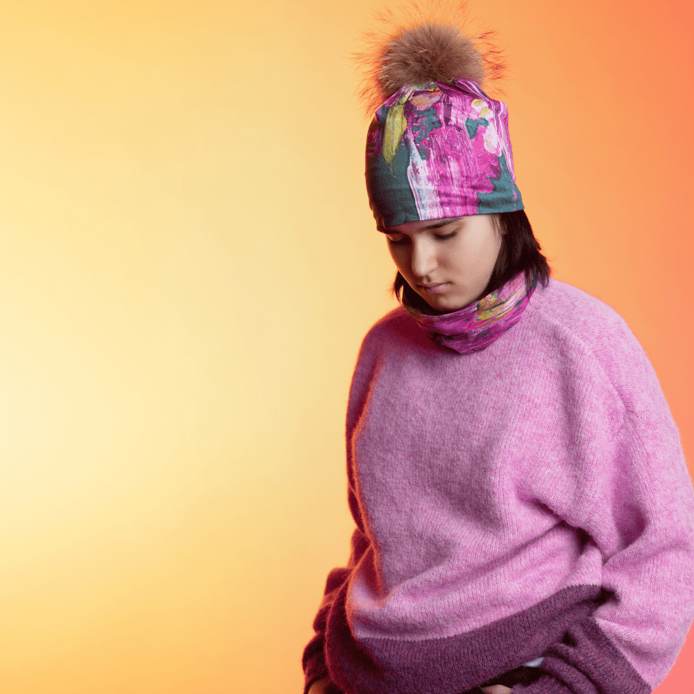 A young person wearing the Flower Glitch Beanie With Pompom and a matching neck gaiter with a fuzzy pink sweater, looking down against a warm gradient background.