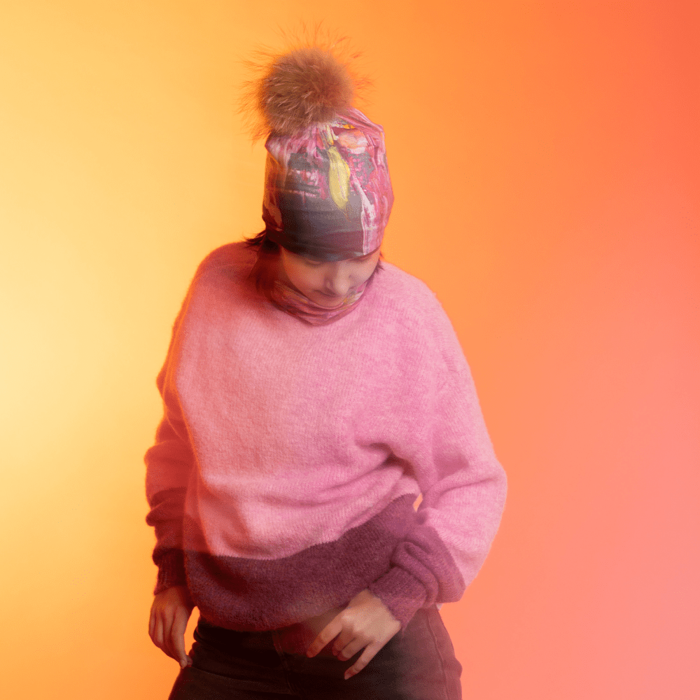 A young person wearing the Flower Glitch Beanie With Pompom and a matching neck gaiter, in motion against a warm gradient background.