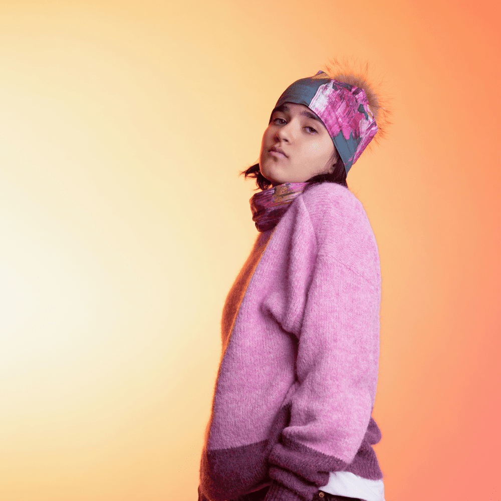 A young person wearing the Flower Glitch Beanie With Pompom and a matching neck gaiter, posed in profile against a warm gradient background.