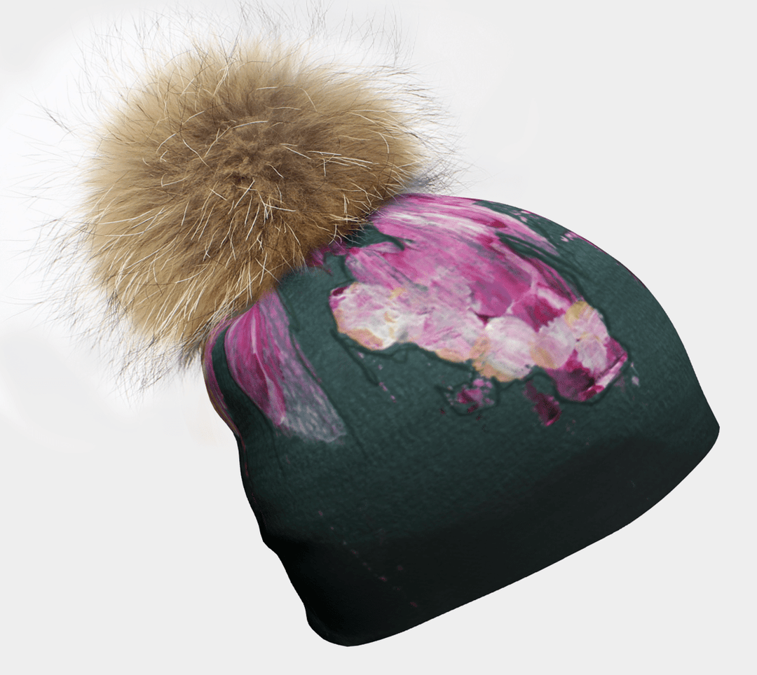 A close-up of the Flower Glitch Beanie With Pompom, showcasing its dark teal base with vibrant pink and white abstract floral brushstrokes and a large, fluffy brown pompom.