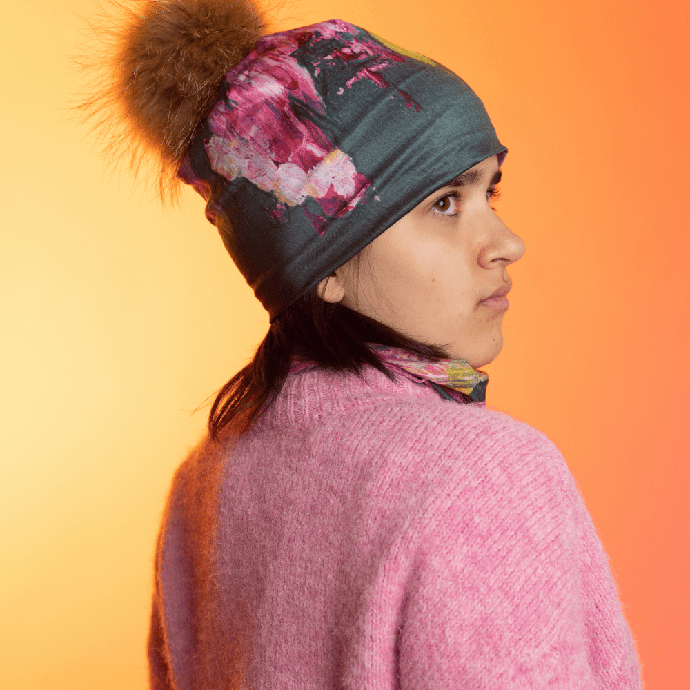 A young person wearing the Flower Glitch Beanie With Pompom and a matching neck gaiter, viewed from the back against a warm gradient background.