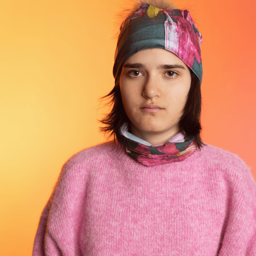 A person wearing a pink sweater and the Flower Glitch neck tube, with a matching beanie on their head, against a gradient background.