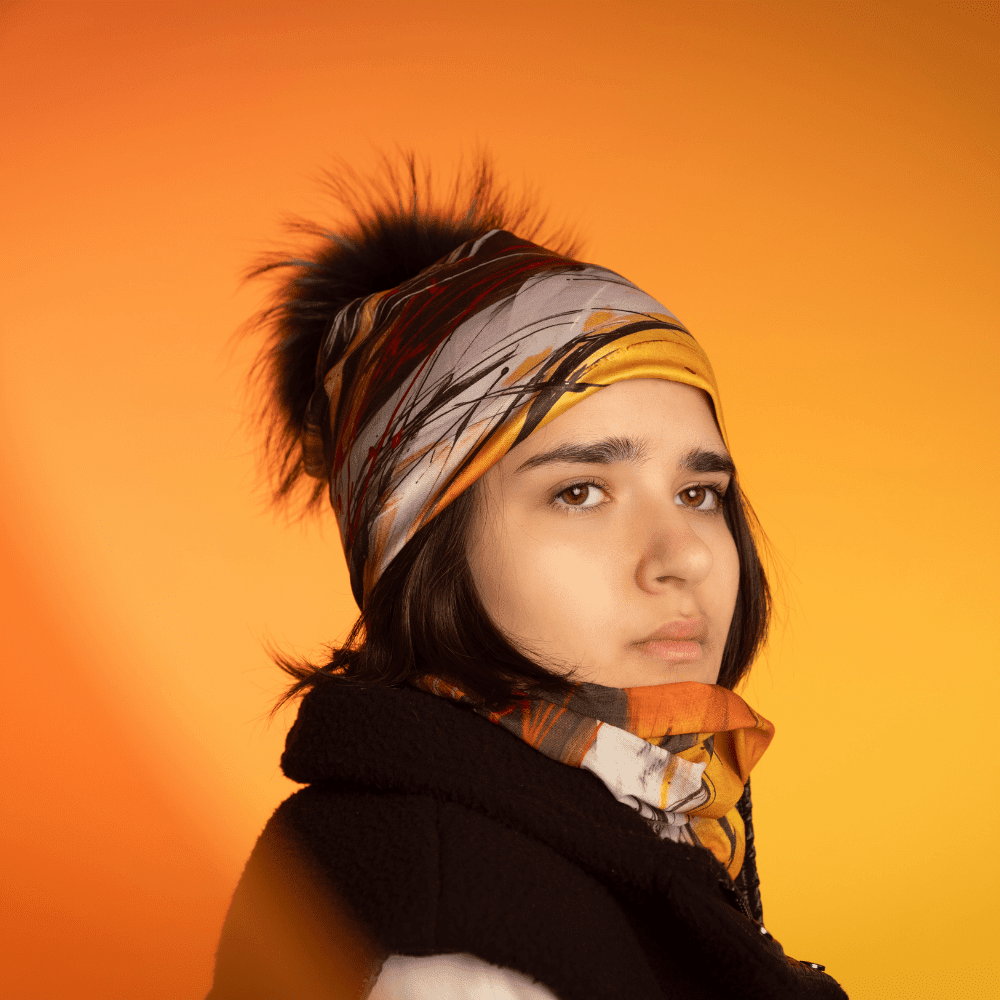 A young woman wears the Le Coeur Beanie with its matching neck gaiter, against a warm orange background.