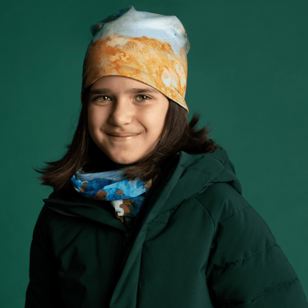 A young girl wearing the Ocean Marble Beanie and matching neck warmer with a dark green winter jacket.