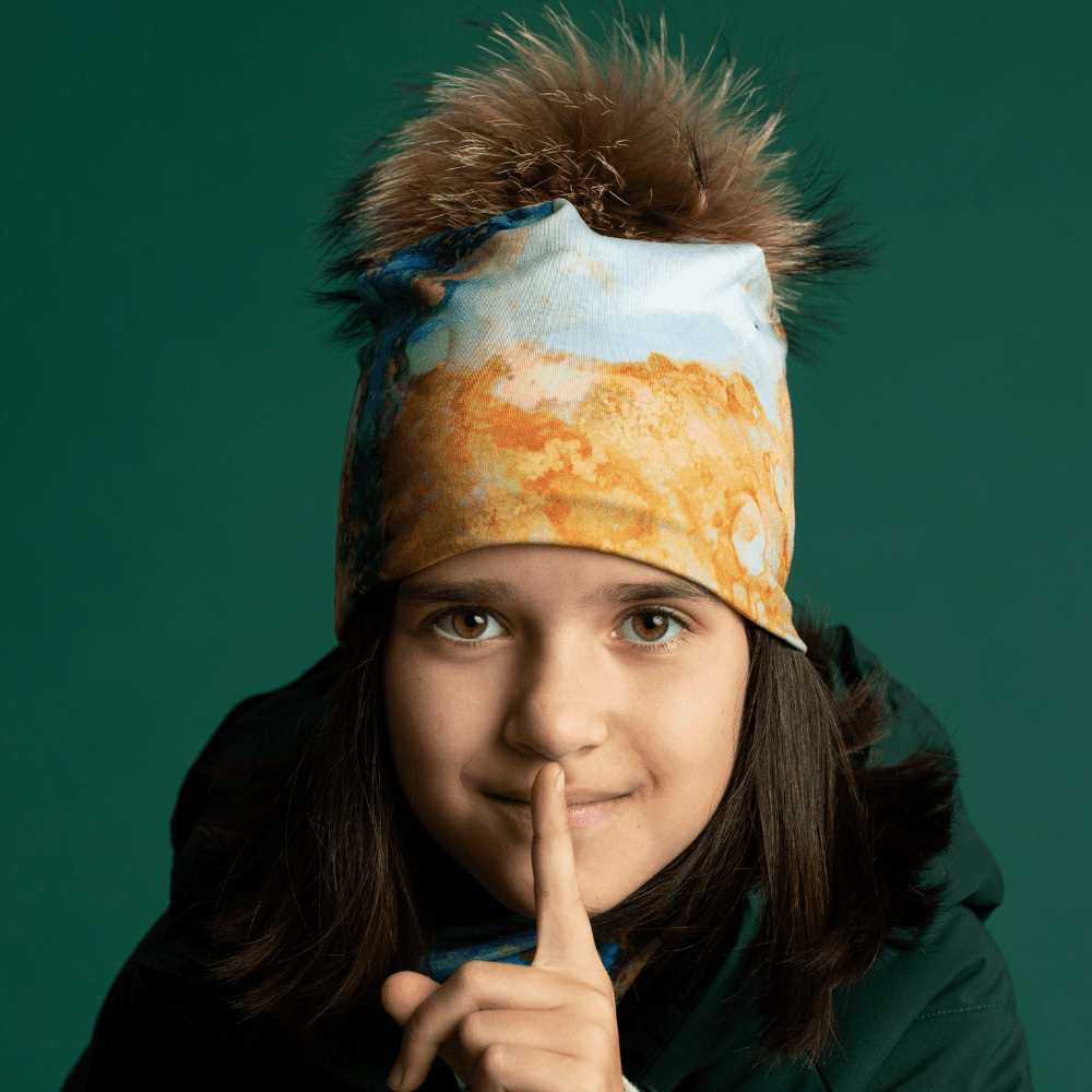 Une jeune fille portant la tuque et la tuque "Marbre Océan" sourit directement à la caméra, le doigt sur ses lèvres.