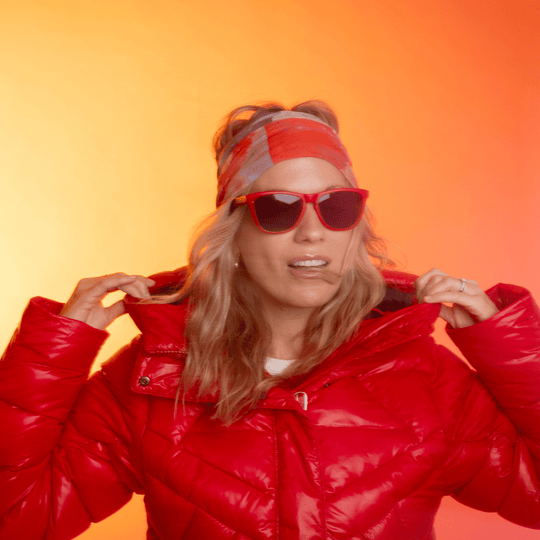 Woman in a red puffer jacket and sunglasses, adjusting her collar, with the Pink &amp; Flames tube as a headband.