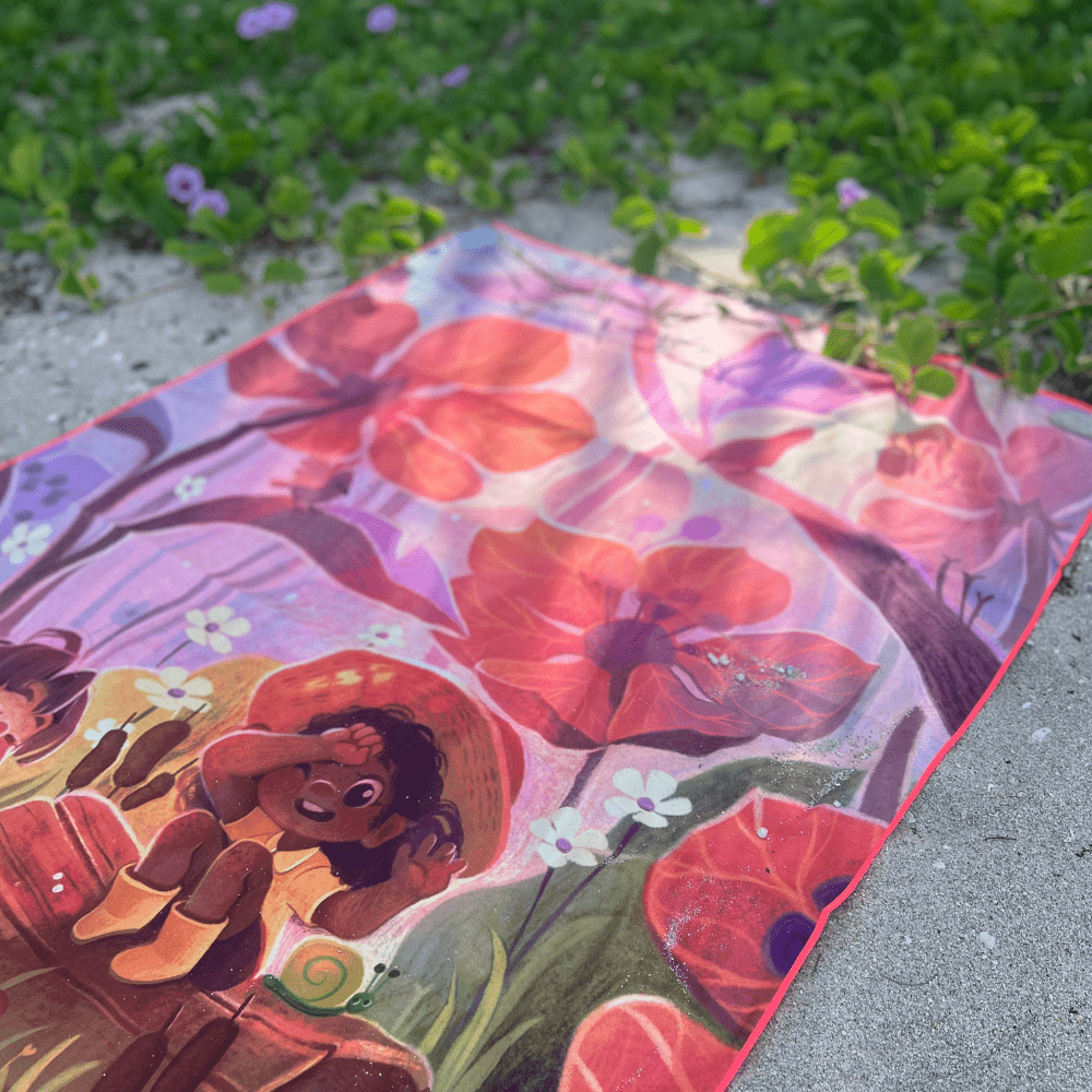 The Playful Rain kids' beach towel spread out on a sandy beach with green foliage.