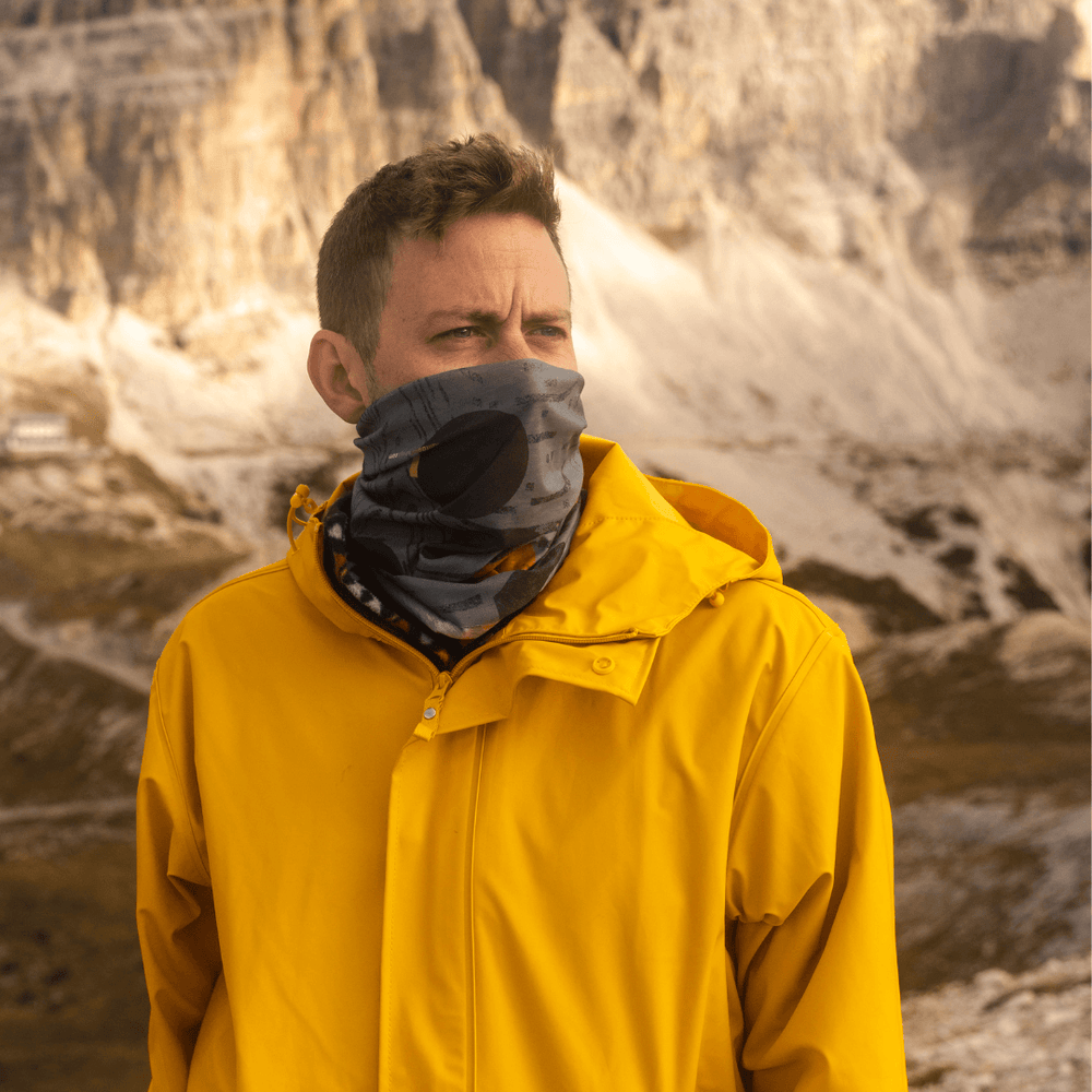 Homme portant une veste jaune avec le tube multifonction Mouvement Orange POLAR comme masque facial dans un décor extérieur montagneux.