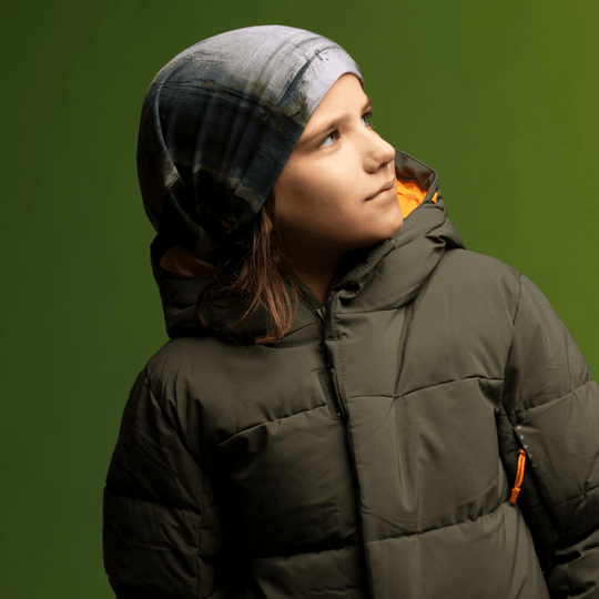 A young person wearing a dark green puffer jacket and a beanie featuring the abstract grey and black "Wall" print, looking upwards against a green background.