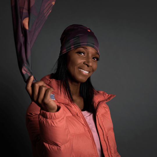 Woman wearing a coral puffer jacket, holding a Purple Geometrics neck tube near her face.