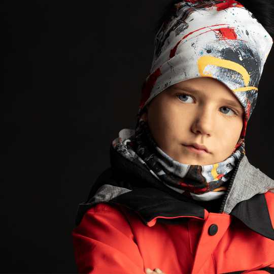 Gros plan sur un jeune garçon portant une tuque et un cache-cou assortis aux motifs abstraits de peinture rouge, noire et jaune sur une veste de ski.