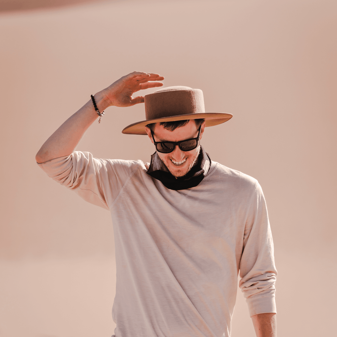 Un homme portant un fedora beige et des lunettes de soleil, saluant de la main avec un tube multifonctionnel noir porté autour de son cou.