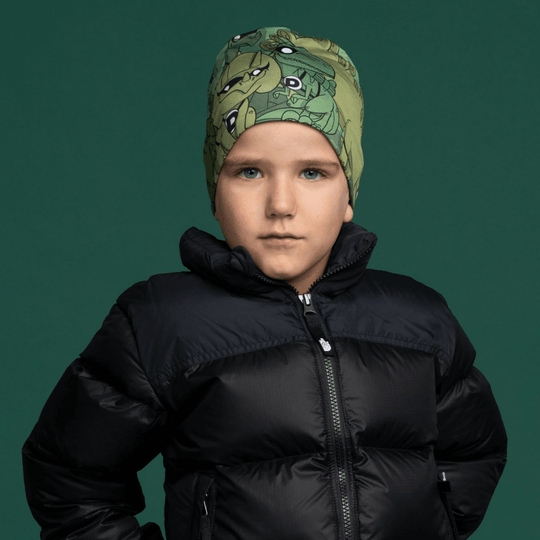 A young boy wearing a green beanie with a dinosaur print, dressed in a black puffer jacket against a dark green background.