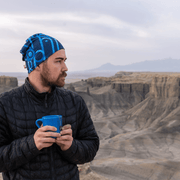 Man in profile drinking coffee and looking into the distance in a canyon landscape. He is wearing the black and blue Mechanic bamboo beanie designed by artist Zaire. Sports cap for men and boys, perfect for soccer, jogging, skiing, skateboarding and hiking.