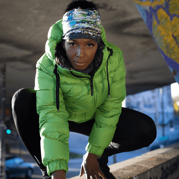 Athletic woman squatting wearing a green winter coat and the Meduse hat designed by Mégane Fortin. This beanie is a beanie with an abstract design and a detachable pom pom