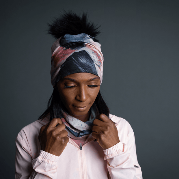 Woman in front with her head down holding her scarf. The woman is wearing a 4 seasons yoga hat designed by Catherine Parent. The hat is colored in pink, grey and beige tones, and has a black pompom. 