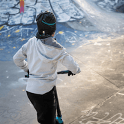 Young boy from behind in a skate park riding a scooter. The young boy is wearing the Robocat hat designed by the artist André Martel