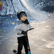 Young boy in a skate park riding a scooter. The young boy is wearing a protective helmet over the Robocat cap designed by the artist André Martel