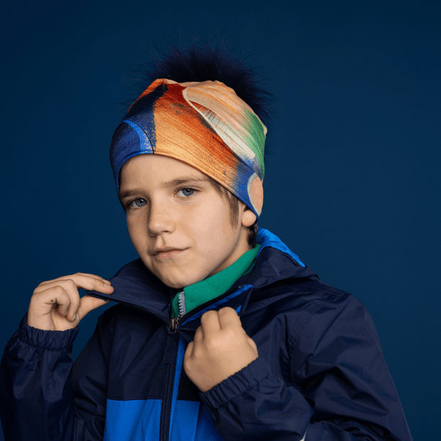Young boy opening his jacket and wearing the yellow and blue cap designed by Anhone. The 4 seasons streetart style beanie has a pretty navy blue pompom.